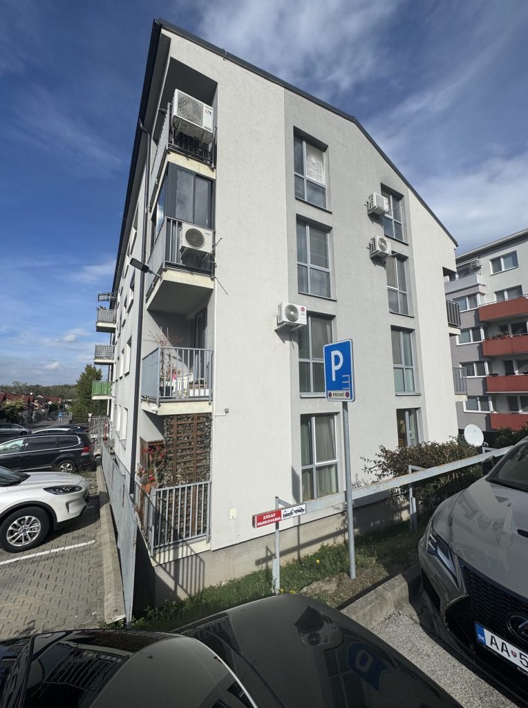 Pohľad zblízka na fasádu bytového domu s balkónmi a zaparkovaným vozidlom v Devínskej Novej Vsi.